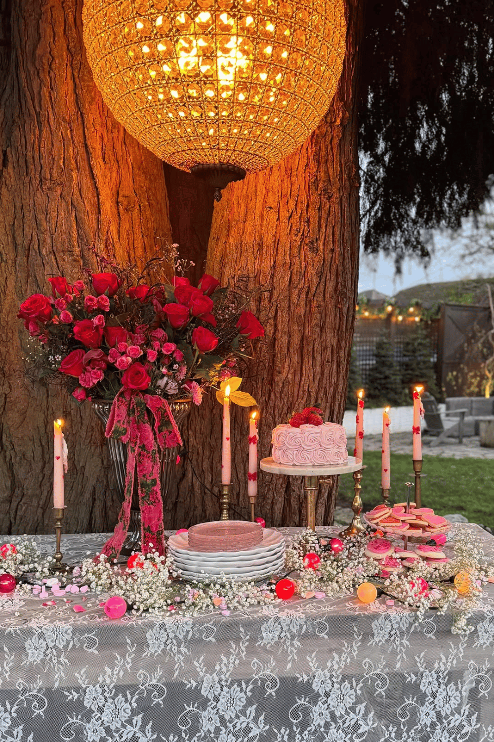 Outdoor Galentine’s dessert table with cake, candles, florals, and lace tablecloth